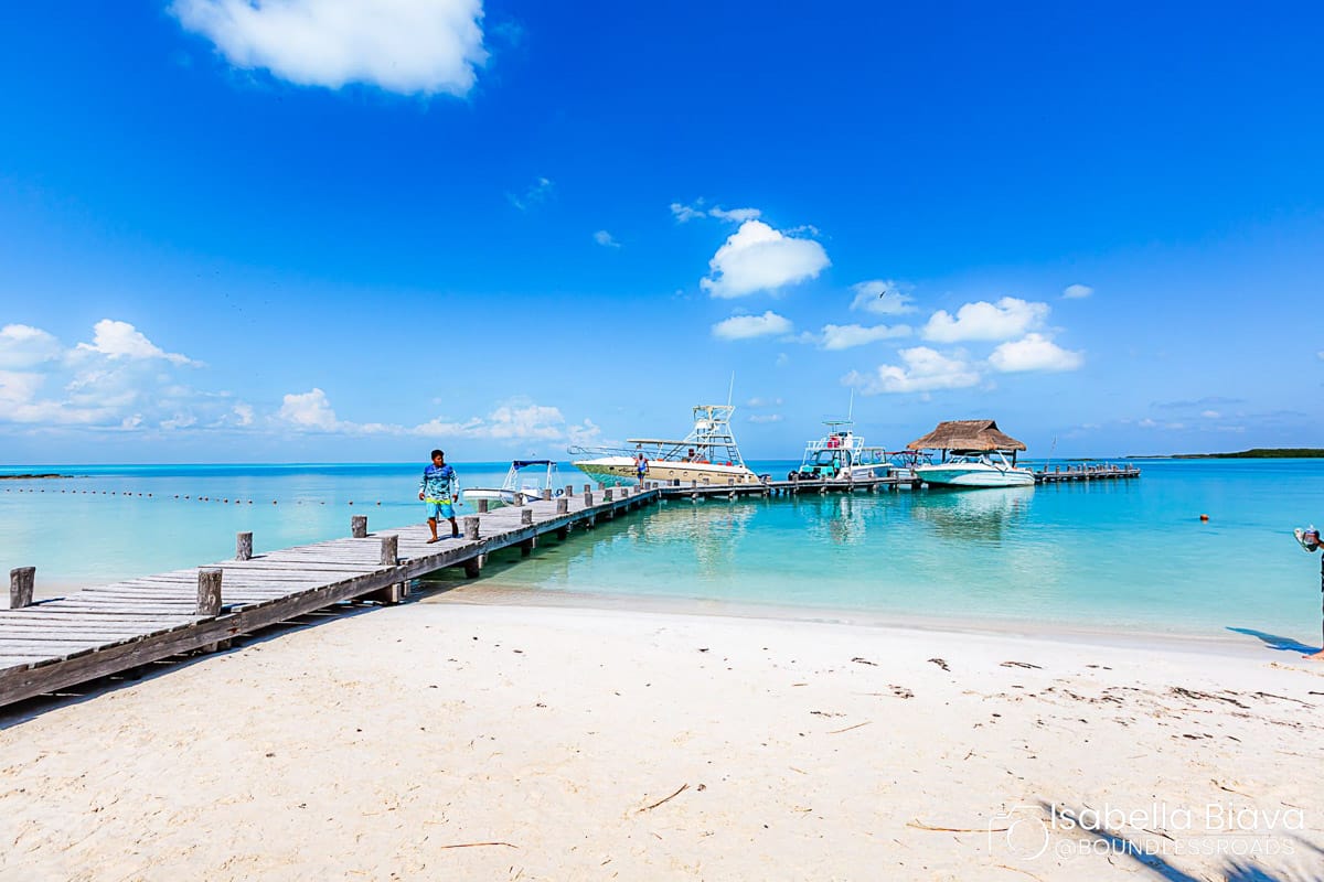 A person walks on a wooden pier extending into crystal-clear water, with boats and blue skies in the background.