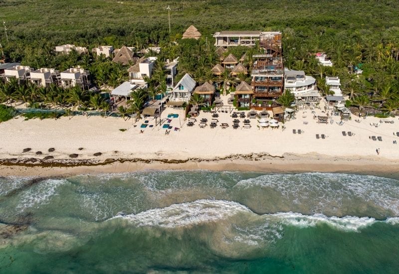 Aerial view of Tulum's coastline, showcasing the turquoise Caribbean Sea meeting white sandy beaches lined with palm trees and beachfront hotels.