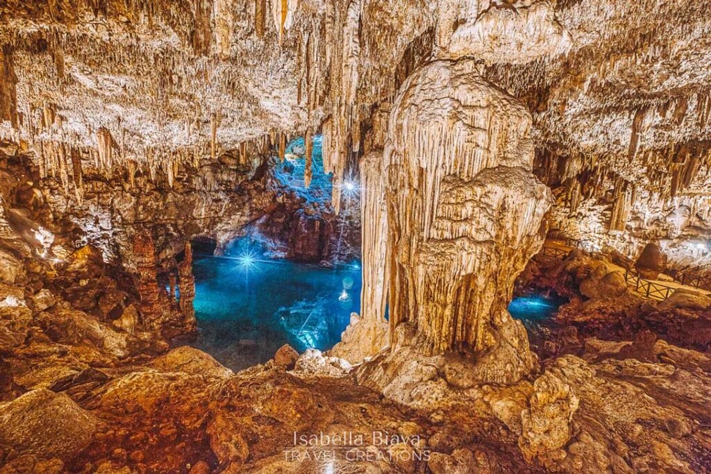 Illuminated cave with stunning stalactites and stalagmites, surrounding a serene underground pool. The warm lighting highlights the intricate rock formations and tranquil blue water.