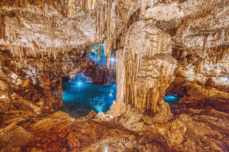 Illuminated cave with stunning stalactites and stalagmites, surrounding a serene underground pool. The warm lighting highlights the intricate rock formations and tranquil blue water.