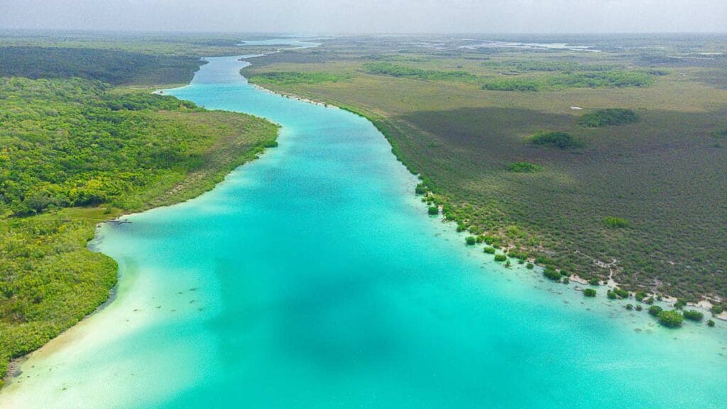 Aerial view of Bacalar Lagoon with vibrant turquoise water, surrounded by lush green vegetation under a hazy sky, creating a serene landscape.