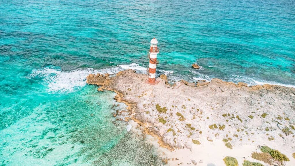 Aerial view of a red and white striped lighthouse on rocky coastline, surrounded by turquoise ocean waters and white waves gently crashing ashore.