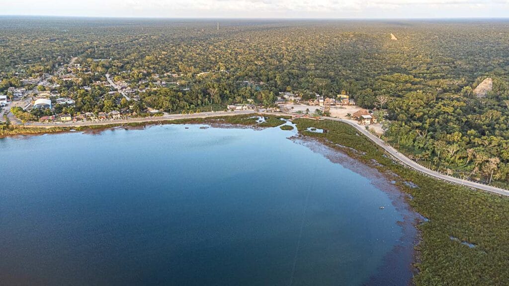 Aerial view of Bacalar Lagoon, surrounded by lush greenery and a small town. A road curves along the waterfront, no visible people.