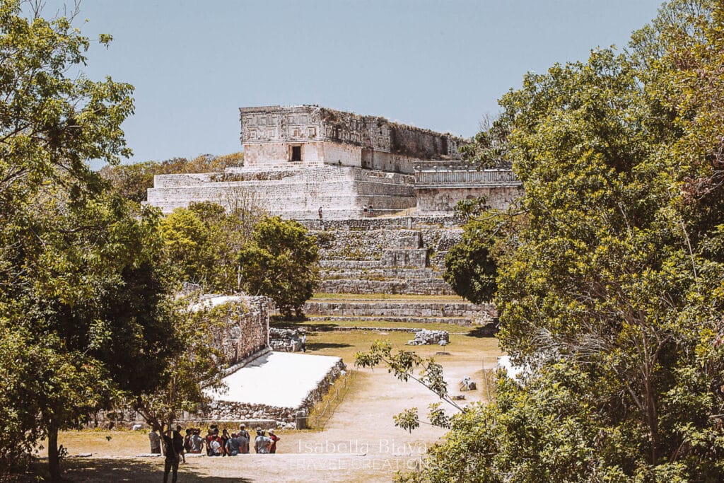 Ancient Mayan ruins surrounded by lush trees under a clear sky, with a group of people gathered at the base, exploring.