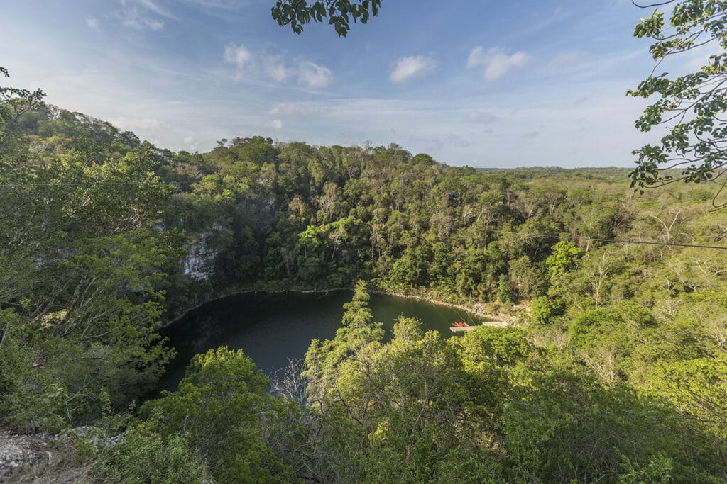 A lush forest surrounds the Sacred Cenote, a circular natural sinkhole with calm water, under a clear sky in a tranquil setting.