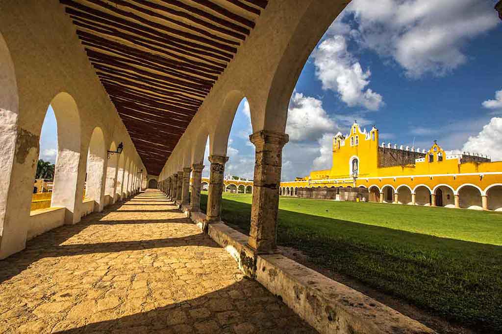 Colonial-style arched walkway leads to the yellow Convent of San Antonio de Padua, framed by lush greenery and a partly cloudy sky.