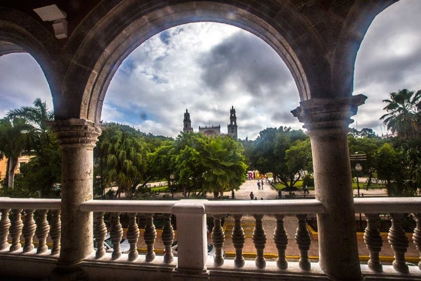 Arched stone balcony overlooks lush park and Mérida Cathedral. Cloudy sky, vibrant greenery, and historic architecture create a serene, picturesque scene.
