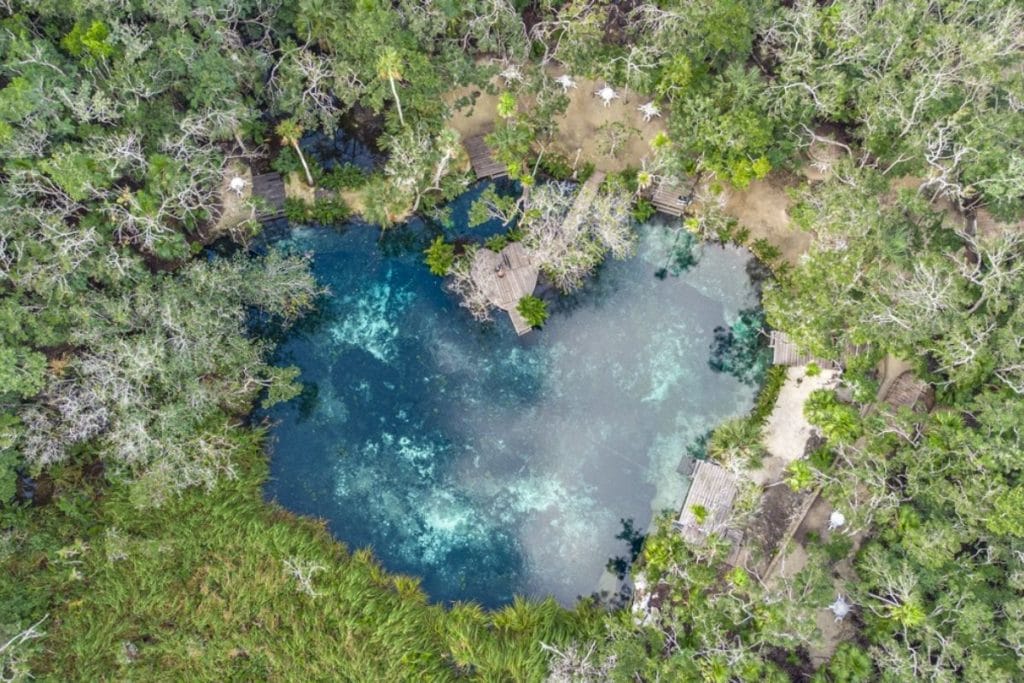 Aerial view of a turquoise, clear cenote surrounded by lush green foliage and a few small structures. People visible around the water's edge.