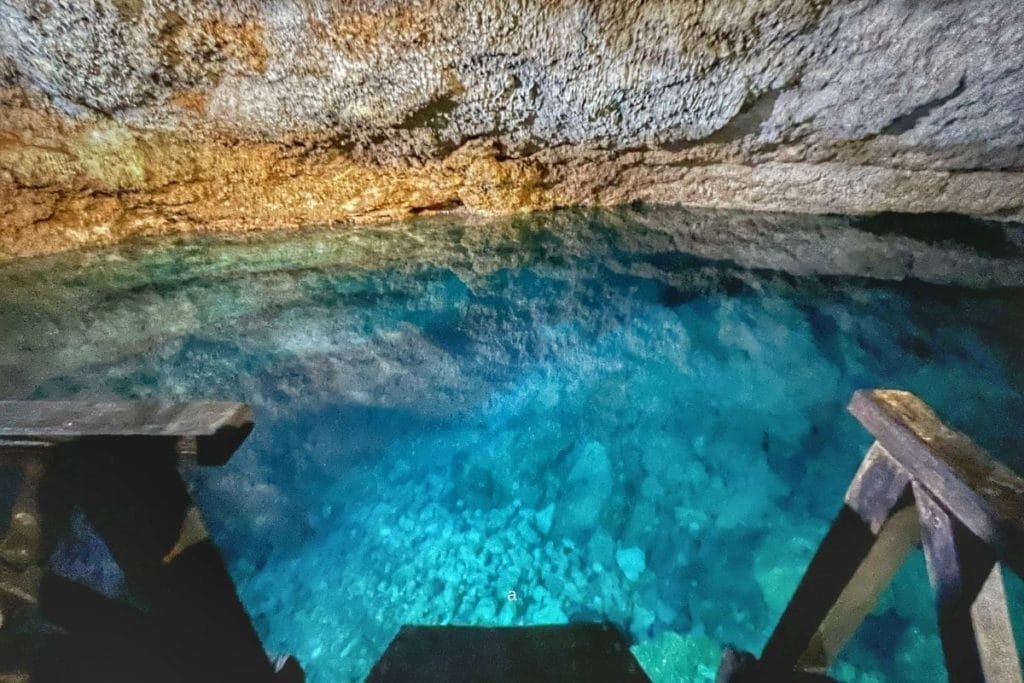 A clear turquoise underground lake inside a rocky cave with wooden stairs leading into the water, illuminated by natural lighting from above.