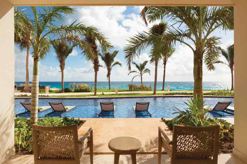  Private patio of a swim-up suite with two chairs facing a tranquil pool, palm trees, and the ocean beyond under a partly cloudy sky.