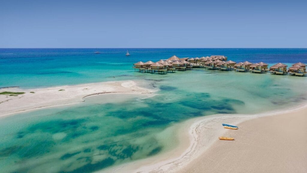 Overwater bungalows extend into the turquoise sea, with sandy beaches and a kayak. A person canoes nearby under a clear blue sky.