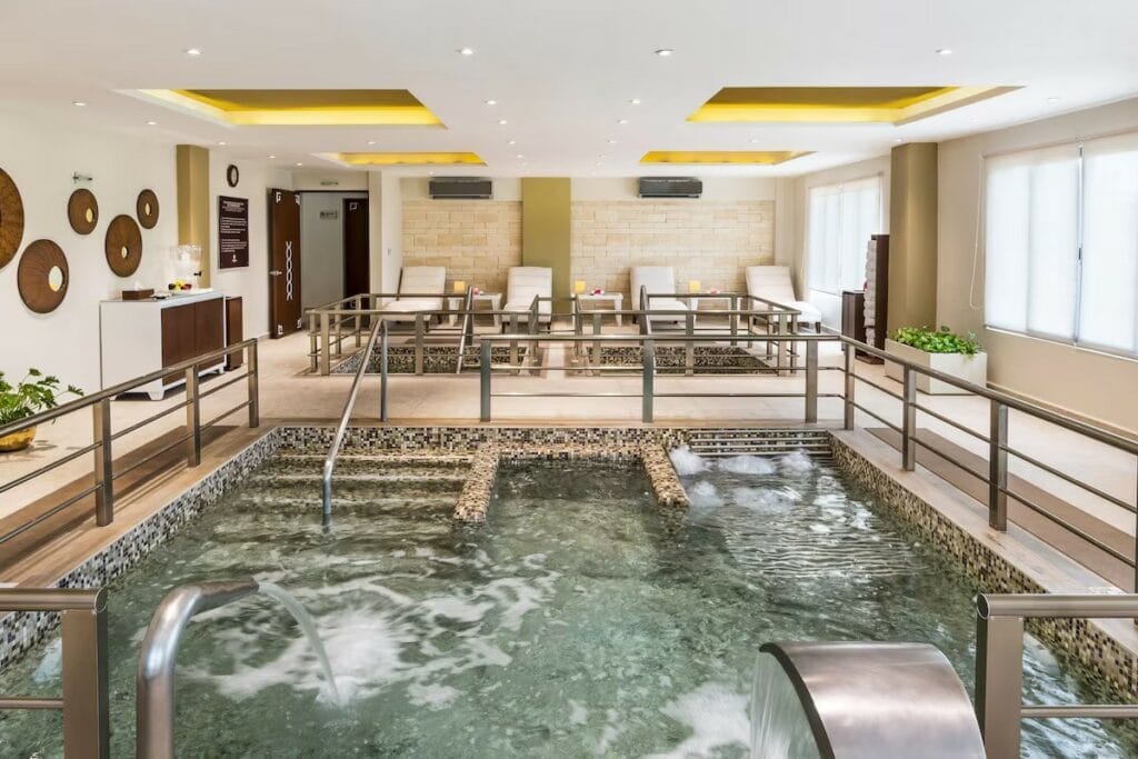  Indoor hydrotherapy spa pool at Palafitos Maroma, with bubbling jets, lounge chairs, and a calming modern design with warm lighting and decorative wall art.

