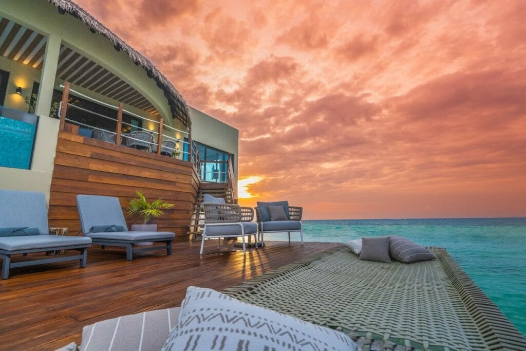 Oceanfront deck at Palafitos Maroma during sunset, with cozy lounge seating and a woven hammock-style net overlooking the water under a vibrant orange and pink sky.

