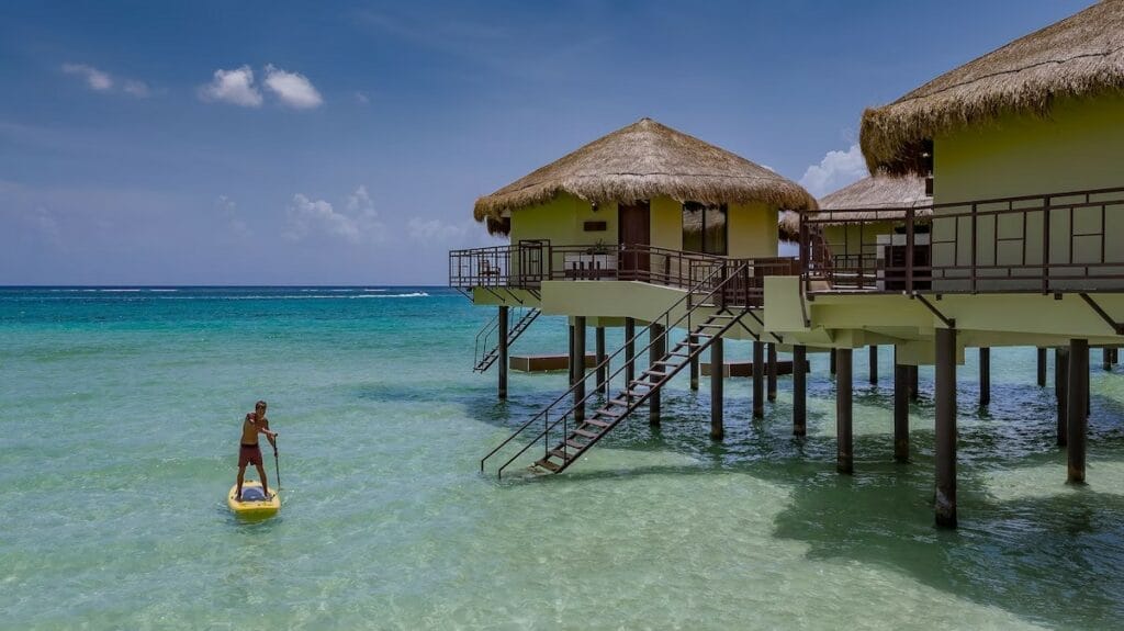 Man paddleboarding in shallow turquoise waters near the stilts of Palafitos overwater villas, with thatched roofs and wooden staircases descending into the sea.

