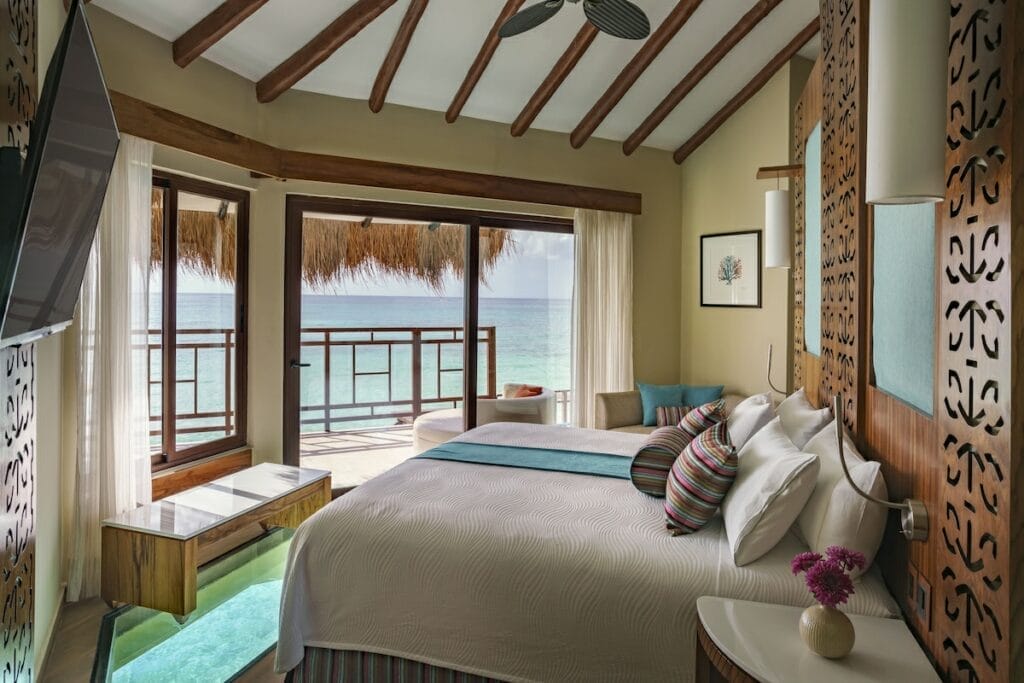 Luxurious overwater bungalow suite with ocean views, featuring a king-sized bed, glass floor panel, thatched roof, and sliding doors opening to a private balcony over the water.
