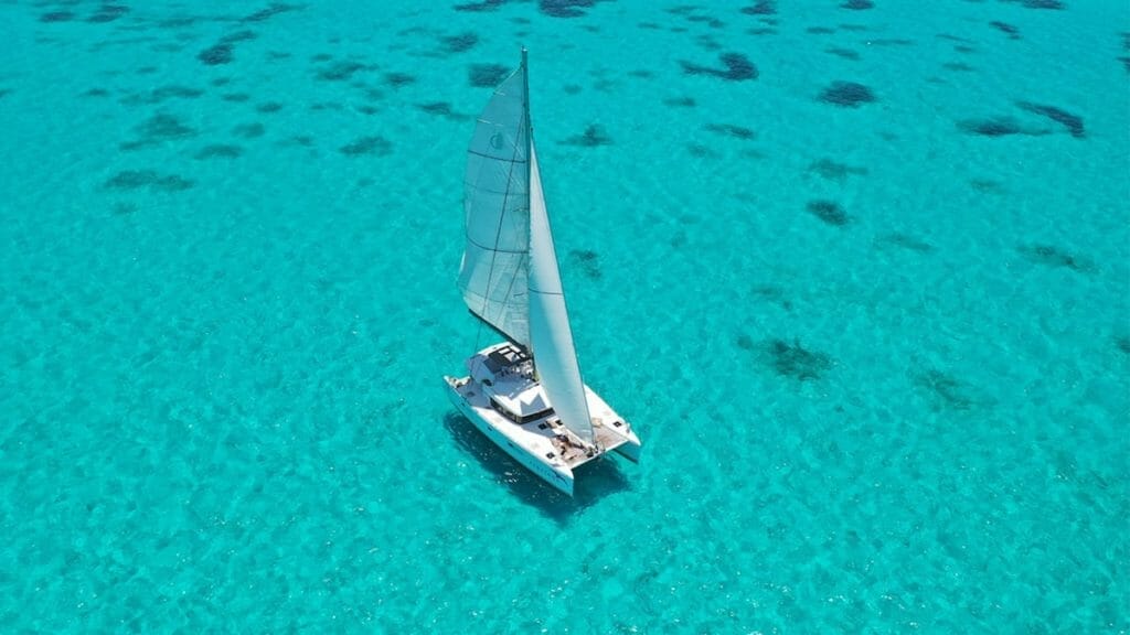 A sailboat glides on clear, turquoise water, surrounded by underwater reefs. The serene scene features vibrant blues and a peaceful, open horizon.