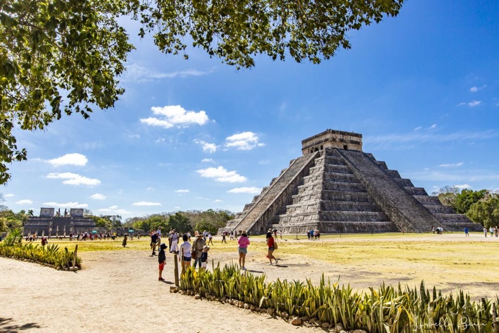 People walking near the ancient Mayan pyramid, El Castillo, at Chichen Itza under a bright blue sky with scattered clouds and lush greenery.