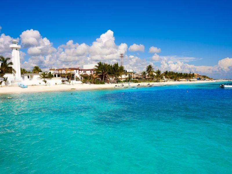 A pristine beach with turquoise water, a lighthouse, and scattered palm trees under a bright blue sky. Small boats dot the shoreline.