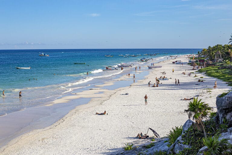 People enjoying a sunny day at a white sandy beach in Tulum. Clear blue waters, boats, and green vegetation enhance the serene coastal scenery.