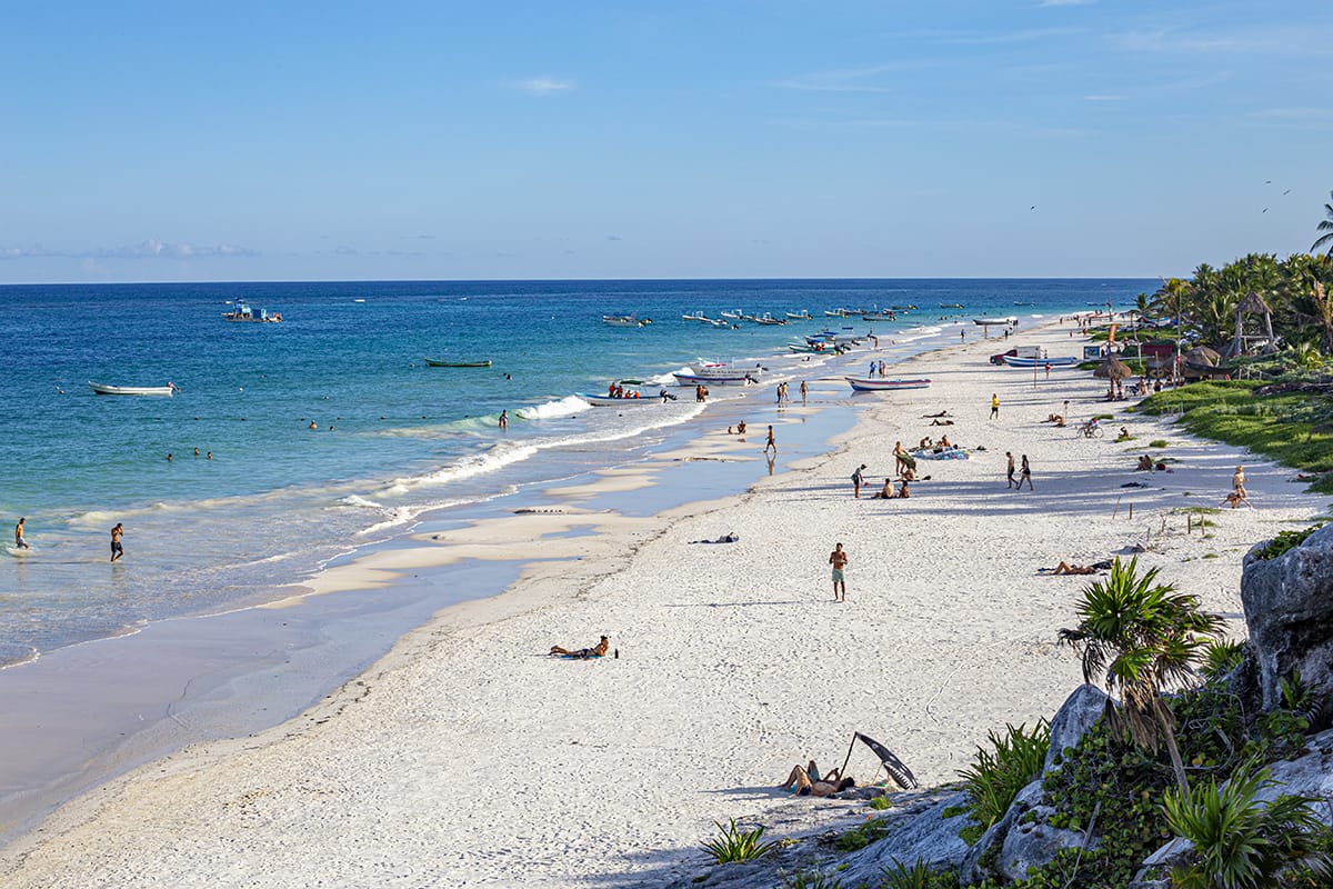 Pristine beach with white sand, clear blue waters, and people relaxing. Palm trees line the shore, while boats float in the distance.