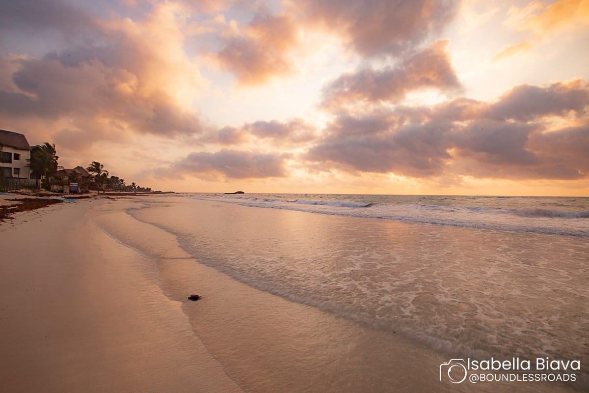 A serene beach at sunrise, with gentle waves and a cloudy sky. Buildings line the sandy shore, creating a tranquil coastal scene.