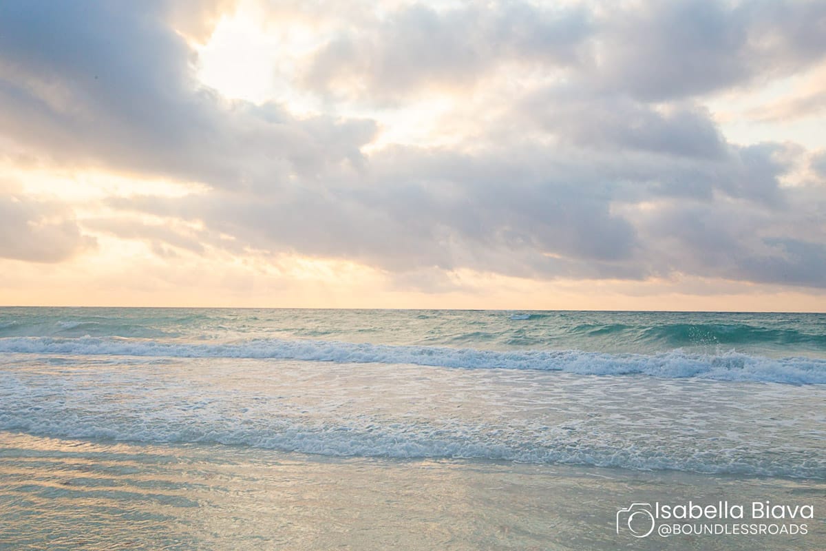 A serene beach view with gentle waves under a partly cloudy sky, illuminated by soft sunlight. No landmarks or people present.