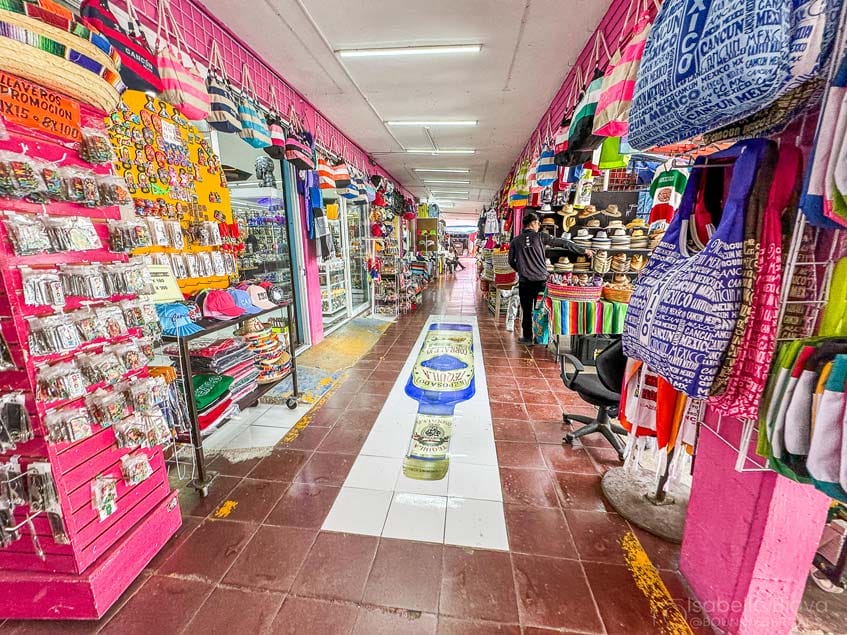 A vibrant indoor market features colorful textiles, hats, and souvenirs. Two people walk through, surrounded by bright pink walls and diverse merchandise displays.