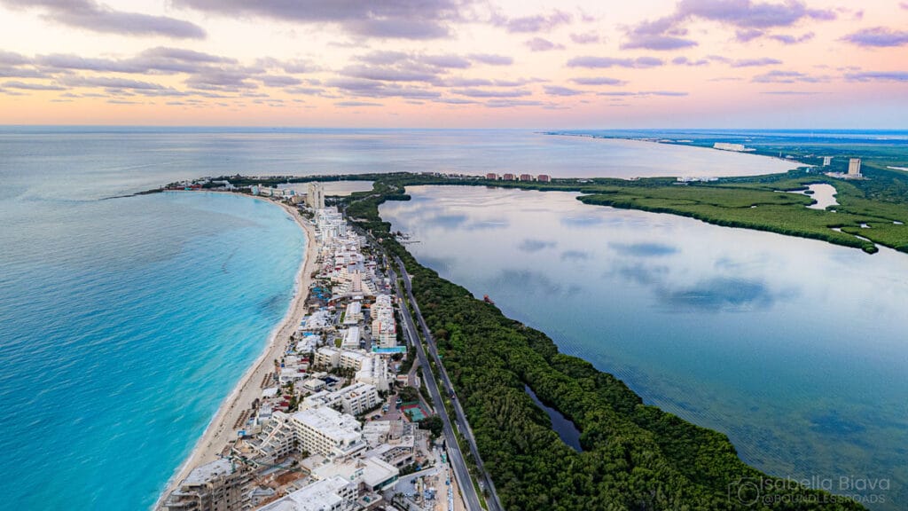 Aerial view of Cancun's coastline, featuring turquoise waters, sandy beaches, resort buildings, green mangroves, and distant lagoon under a colorful sky.