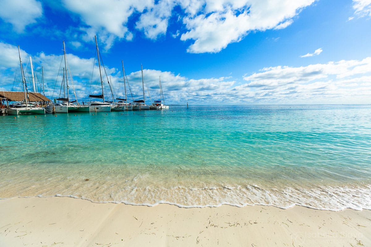 Tranquil beach scene with clear blue water, sandy shore, and docked sailboats under a partly cloudy sky. No landmarks or historical buildings visible.