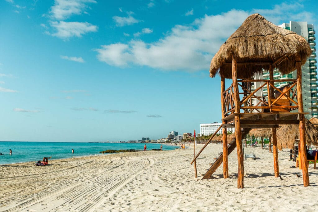 A sandy beach with a thatched lifeguard tower, people by the shore, and modern buildings in the background under a clear, blue sky.