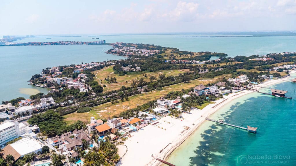 Aerial view of sandy beaches and houses on Isla Mujeres, surrounded by clear blue waters and lush greenery under a partly cloudy sky.