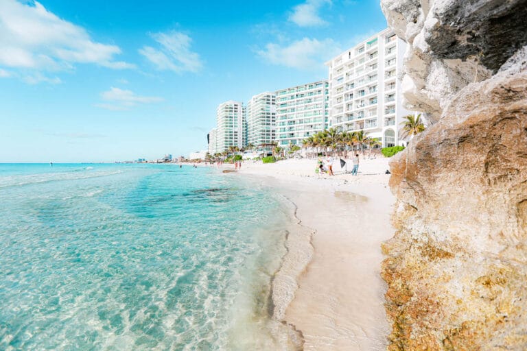 Clear turquoise waters lap against a sandy beach lined with people, palm trees, and several modern buildings under a bright blue sky.