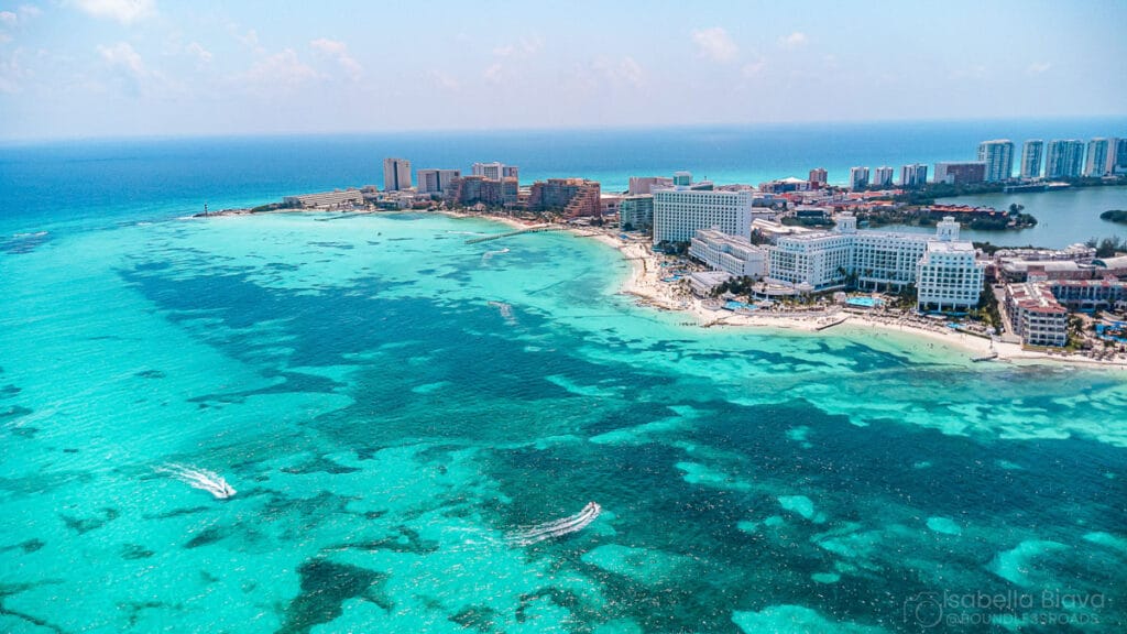Aerial view of Cancun, featuring clear turquoise waters, hotels, and a vibrant coastline under a sunny sky. Boats glide on the tranquil sea.