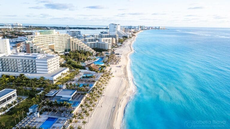 Aerial view of Cancun beaches, hotels, and turquoise waters; palm trees line the shore with distant skyline under a clear sky.