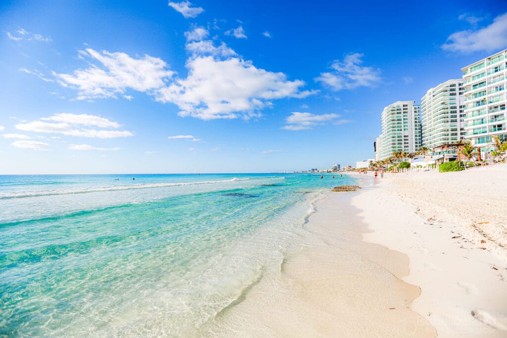 A picturesque beach scene with clear turquoise water, white sand, and modern buildings lining the shoreline under a bright blue sky.