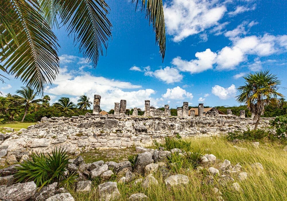 Ancient ruins of the Tulum archaeological site under a bright blue sky, bordered by palm trees and grass, showcasing historical Maya architecture.