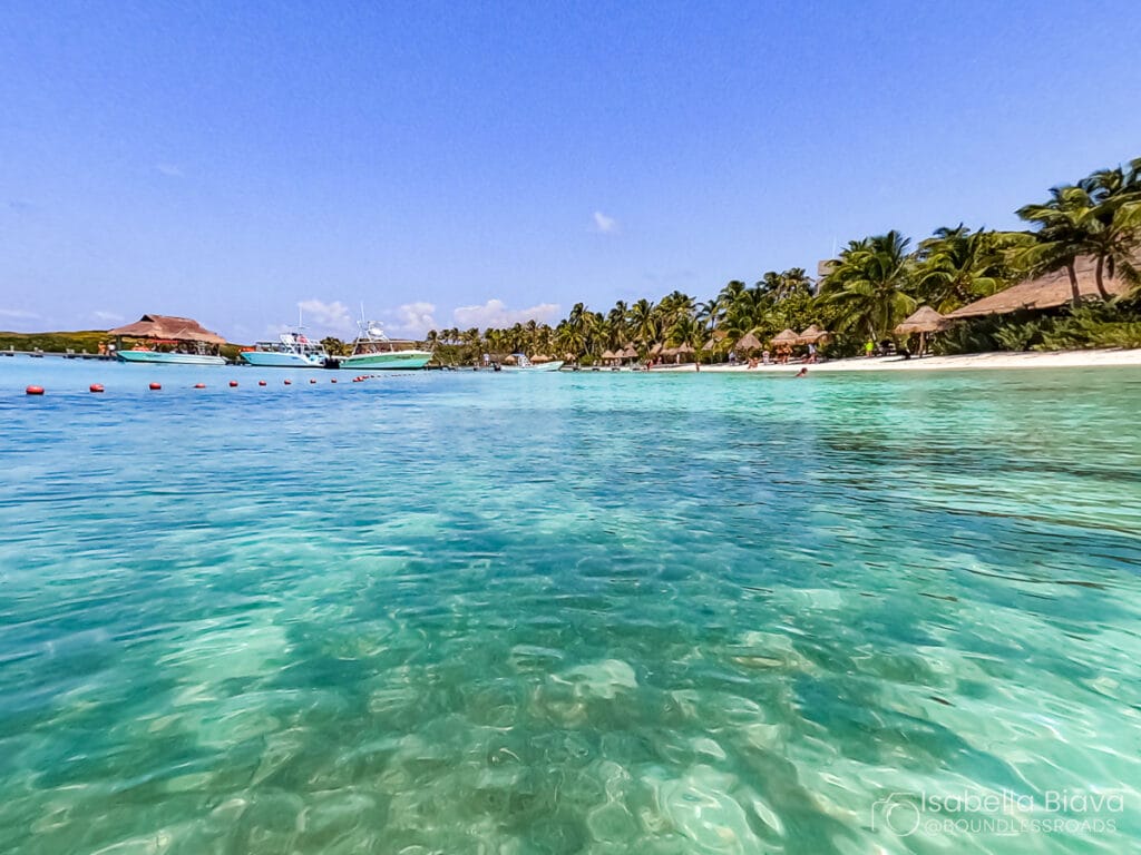 Clear turquoise water with a white sandy beach, palm trees, and people in the distance. Boats are anchored nearby under a sunny sky.