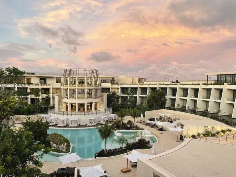 A luxurious resort with a central pavilion and pool, surrounded by modern architecture, under a beautiful sunset sky, creating a serene atmosphere.