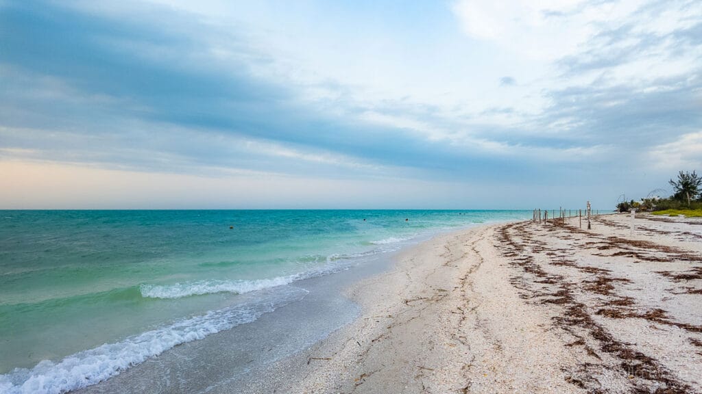 Sandy beach with turquoise waves under a cloudy sky, bordered by a simple fence along the shoreline, creating a serene coastal scene.