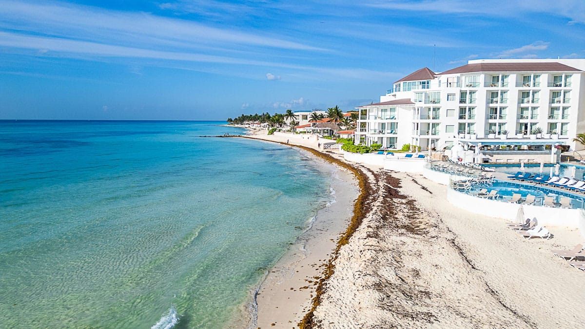 A beachfront resort overlooks clear, turquoise ocean waters with lounge chairs on a sandy shore under a blue sky, near scattered palm trees.