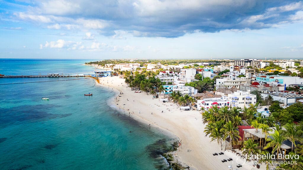 Aerial view of a coastal town with sandy beach, turquoise sea, palm trees, and people along the shore under partly cloudy skies.