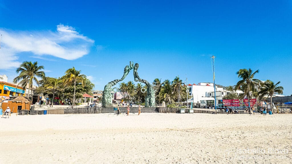 Beach scene featuring Portal Maya sculpture, surrounded by palm trees and buildings, with people walking on sandy shore under a bright blue sky.