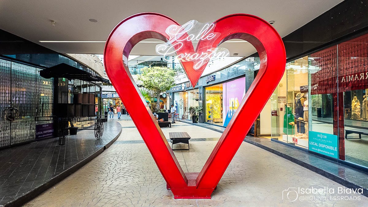 A red heart sculpture stands in a shopping arcade, with neon text above. The area is lined with shops and a tree.