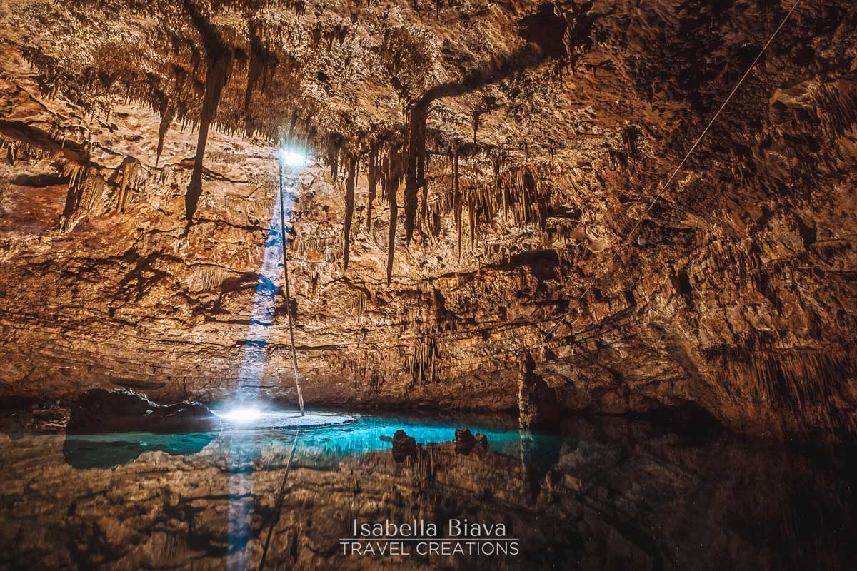 A naturally lit cave with stalactites reflected in a tranquil pool of water, creating a serene and mystical underground scene.