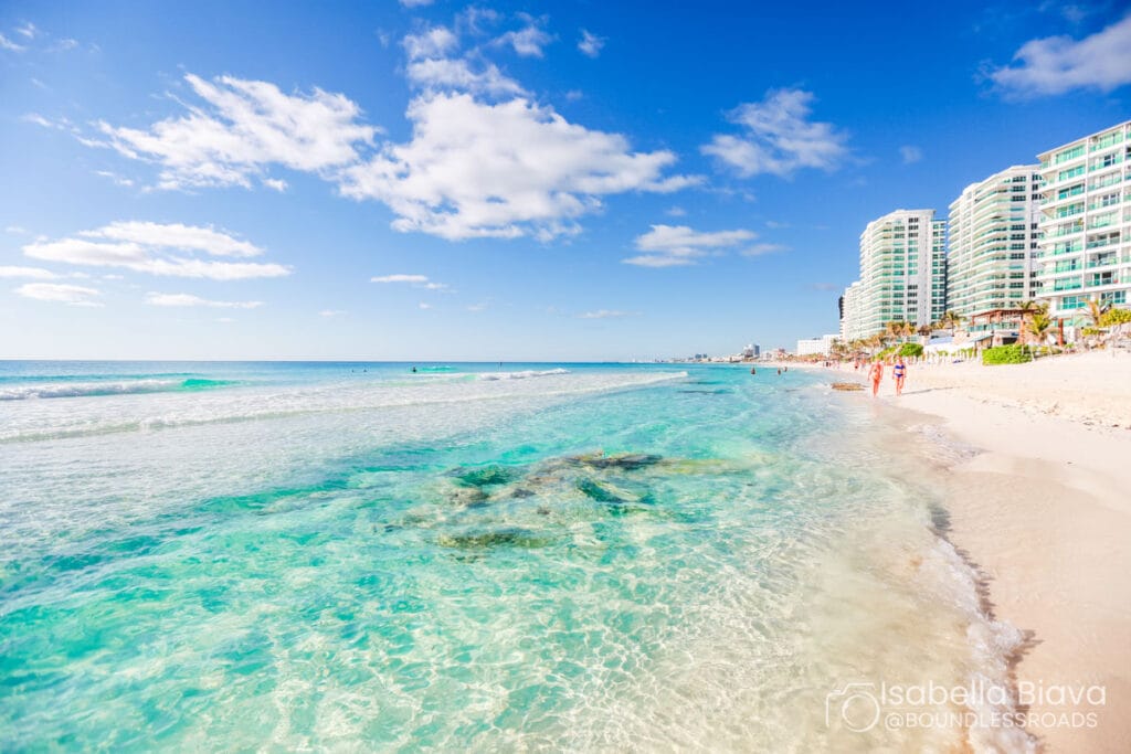 A beautiful beach scene in Cancun with clear turquoise waters, white sand, and tall modern buildings along the coastline under a bright blue sky.