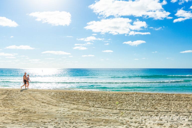 Two people walk along a sunny beach with clear skies and gentle waves, enjoying a tranquil seaside setting under bright, fluffy clouds.