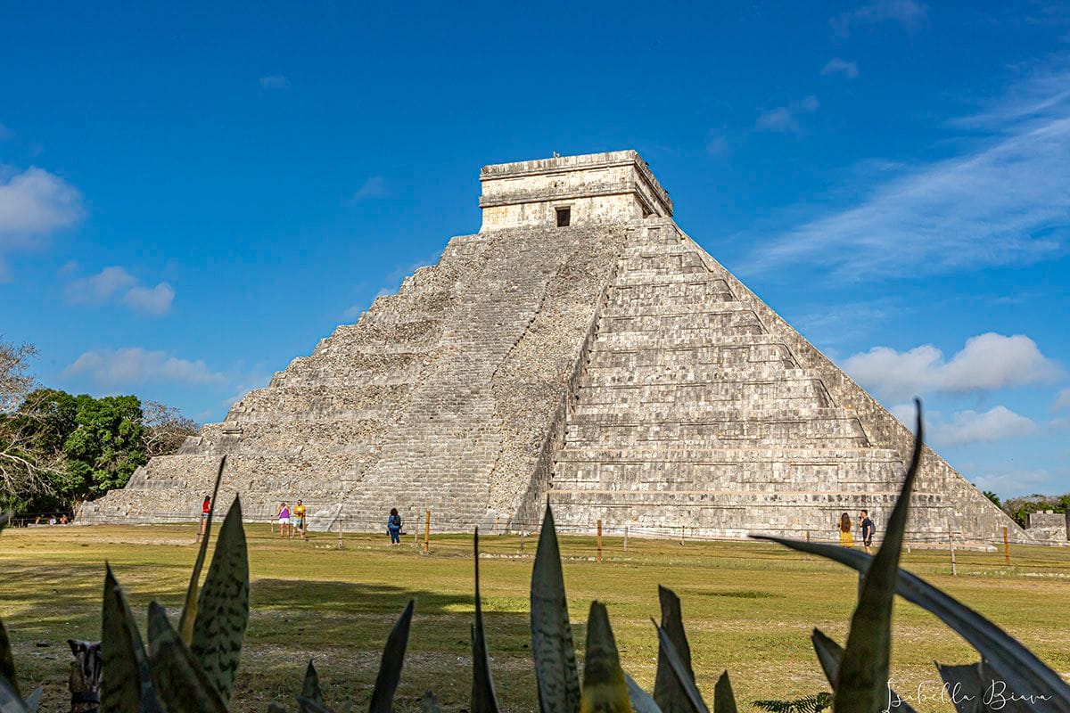 A large pyramid structure, El Castillo at Chichen Itza, stands under a blue sky with several people walking nearby, surrounded by greenery.