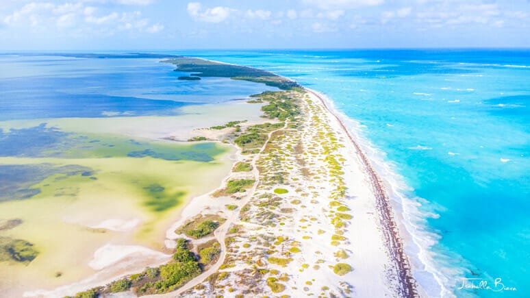 Aerial view of a pristine beach with turquoise waters and sandy shoreline, bordered by lush greenery under a blue sky with scattered clouds.