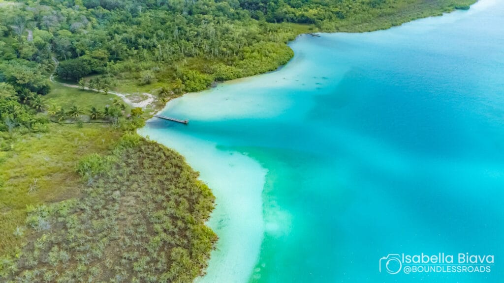 Aerial view of crystal-clear turquoise lagoon surrounded by lush, green forest and a wooden pier extending into the water. No buildings visible.