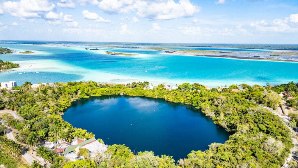 Aerial view of Cenote Azul surrounded by lush greenery, adjacent to turquoise waters of Lake Bacalar under a clear blue sky with scattered clouds.
